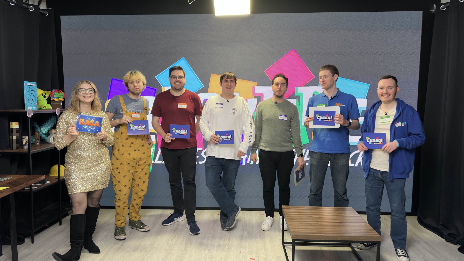 Chelsea, Stuart, Adam, Stuart, George, Bez, and Callum holding RASA Studios and Cubed! branded cue cards, standing in front of a wall-spanning LED screen displaying the logo of Quickfire.