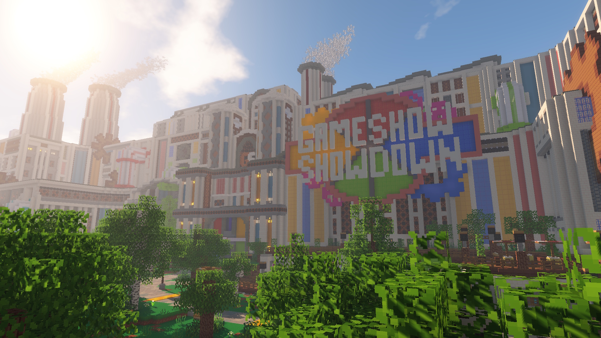 A screenshot within Minecraft of the Gameshow Showdown Factory. The logo of Gameshow Showdown is shown many metres tall on the wall of the factory. In front of the factory are a decking area with statues, and a number of trees. The walls of the factory have large pipes coming out of them in various places, and the walls themselves are composed of different panels, many of which are blue and yellow. There are large chimneys on the roof with smoke coming out of them.