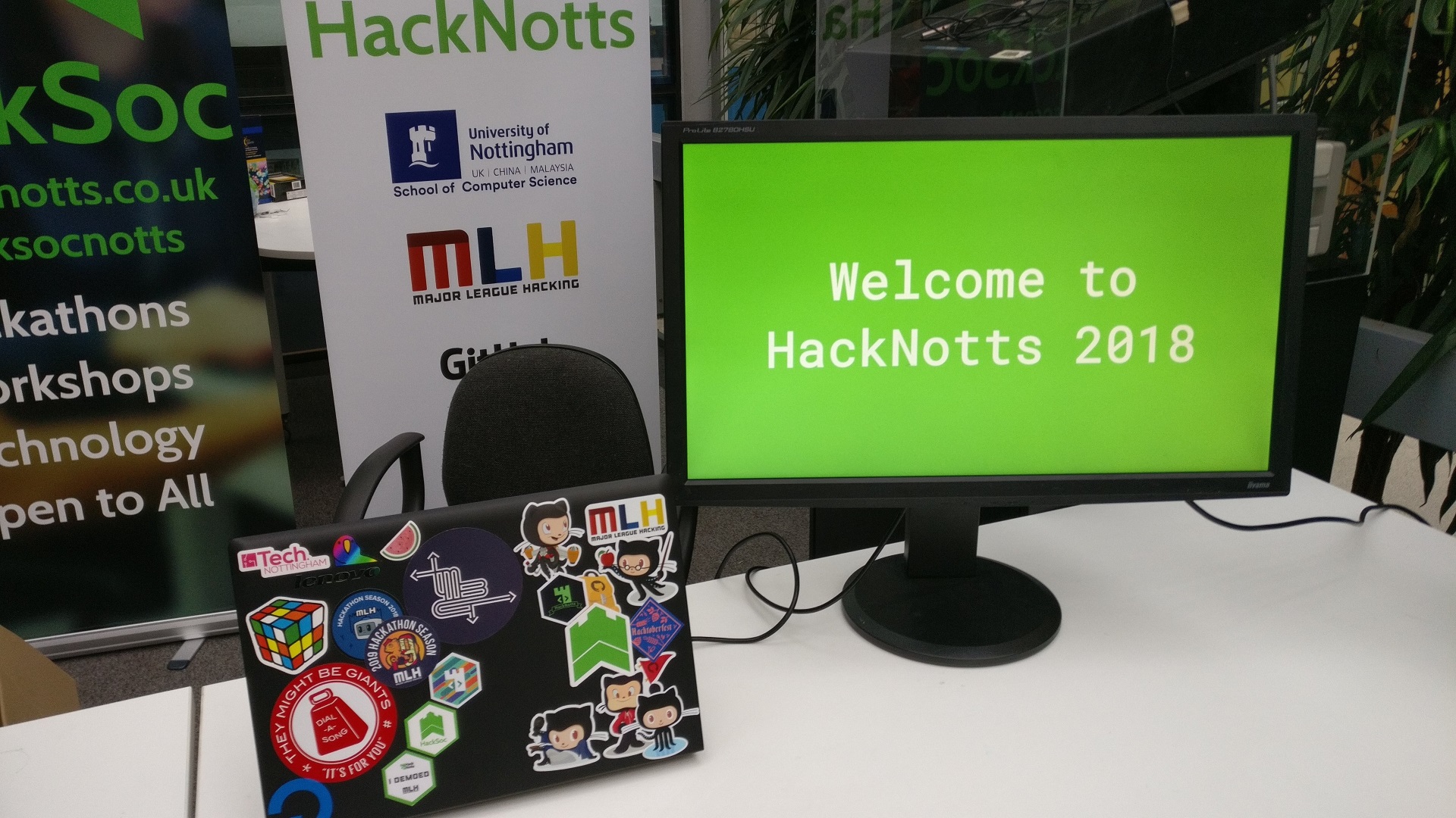 A laptop and a monitor are sat on a desk. The back of the laptop can be seen, with various programming-related stickers on the back. The monitor is facing the viewer, and on it is the text "Welcome to HackNotts 2018". In the background, some banners for HackSoc and HackNotts can be seen.