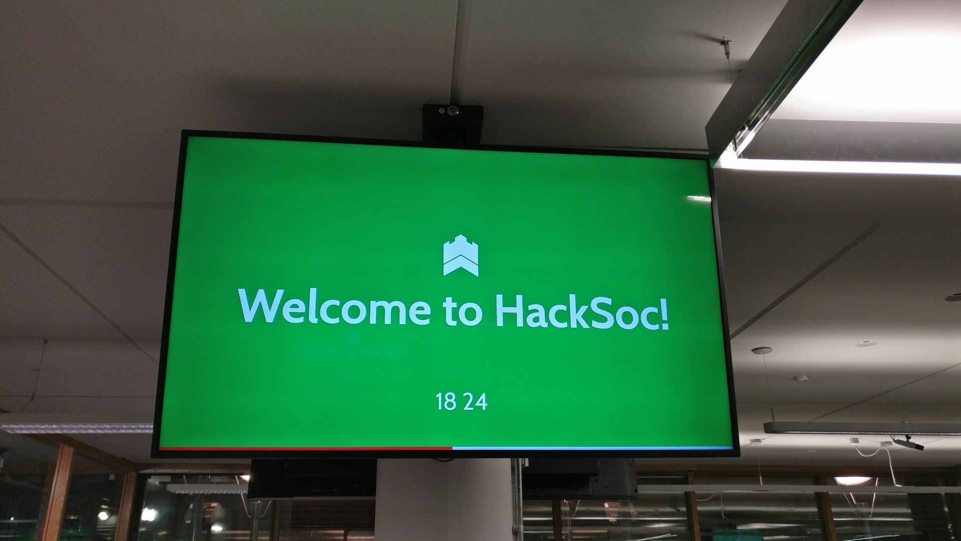 A television mounted on a pillar shows the text "Welcome to HackSoc!", with the time 18:24 visible in small text below it. The HackSoc logo is shown above the main text.