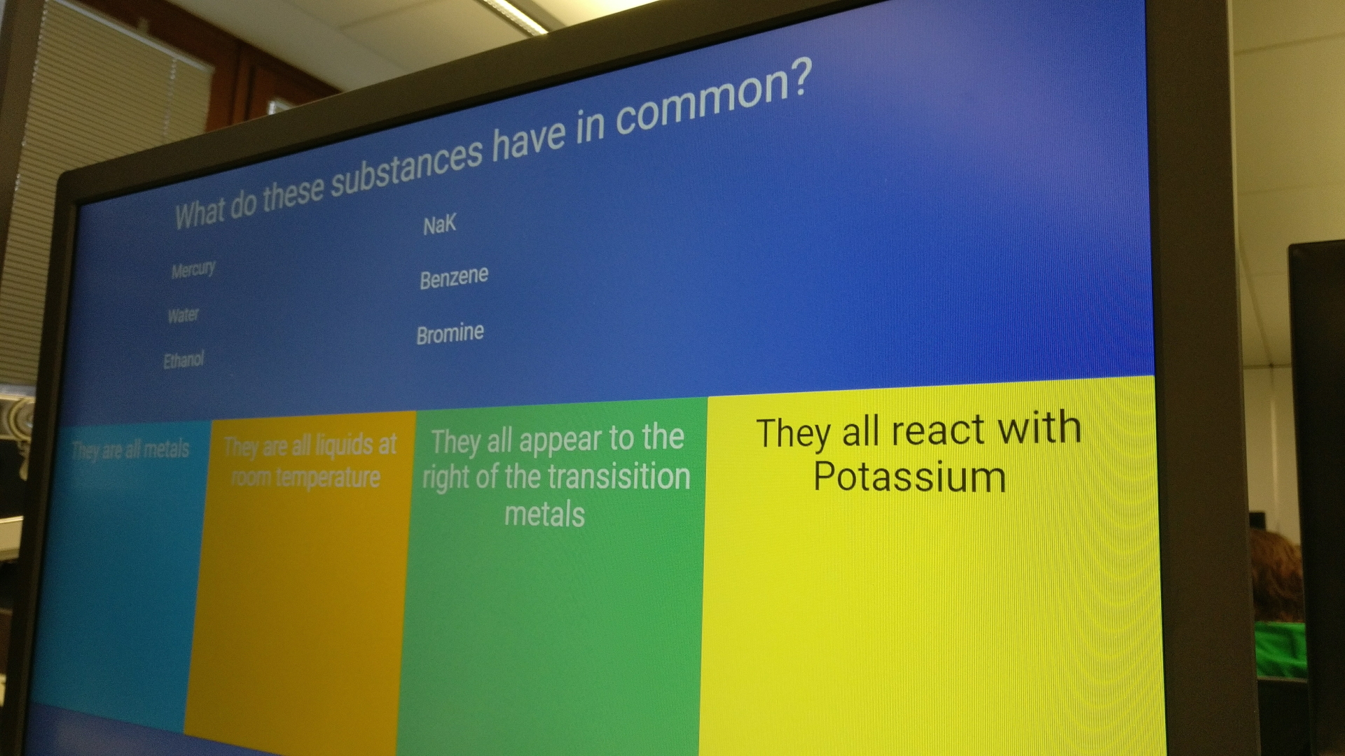 A photograph of a computer monitor. On the screen a question can be seen, reading "What do these substances have in common? Mercury, Water, Ethanol, NaK, Benzene, Bromine." At the bottom of the screen are four answers, each with a different coloured background. They read "They are all metals", "They are all liquids at room temperature", "They all appear to the right of the transition metals", and "They all react with Potassium".