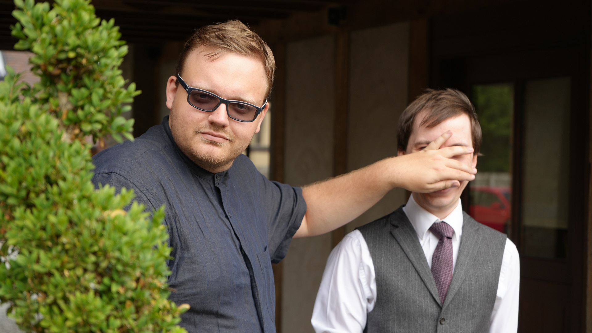 A photo of Ben and Conrad stood outside in front of a wall next to a tall green plant, looking directly at the camera. Ben is wearing a dark blue, short sleeved shirt and slightly tinted glasses. Conrad is stood to his left and smiling at the camera, wearing a white shirt, purple tie, and gray waistcoat. Ben's left hand is reaching out and covering almost all of Conrad's face.