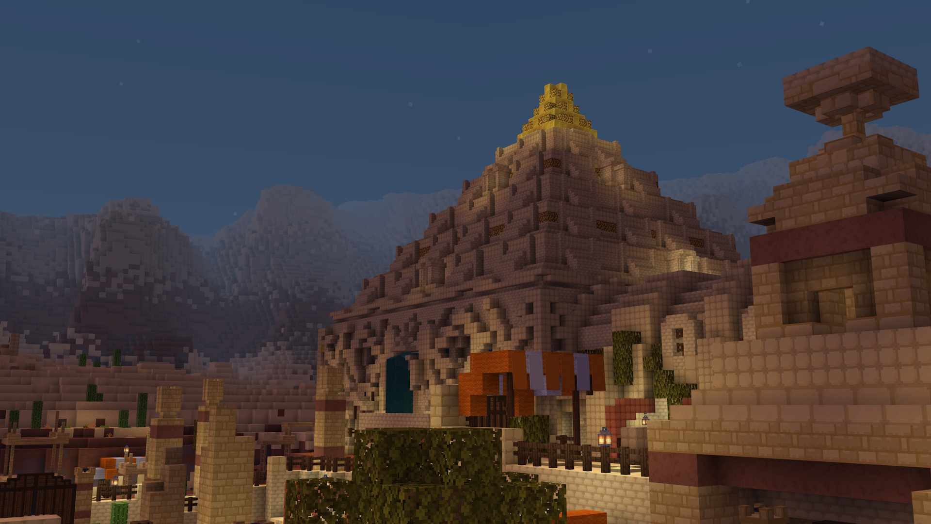 A screenshot within Minecraft of the game arena in Skeleton Survivors. The focus of the image is the large sandstone pyramid in the background. Closer to the camera on the right is an abstract statue made out of sandstone. In the bottom middle of the image, there's the top of a tree. Nearby, there are some orange and white stripy canopies. At the back left, there's a large wooden door that is closed. In the far distance, large mountains surround the area.